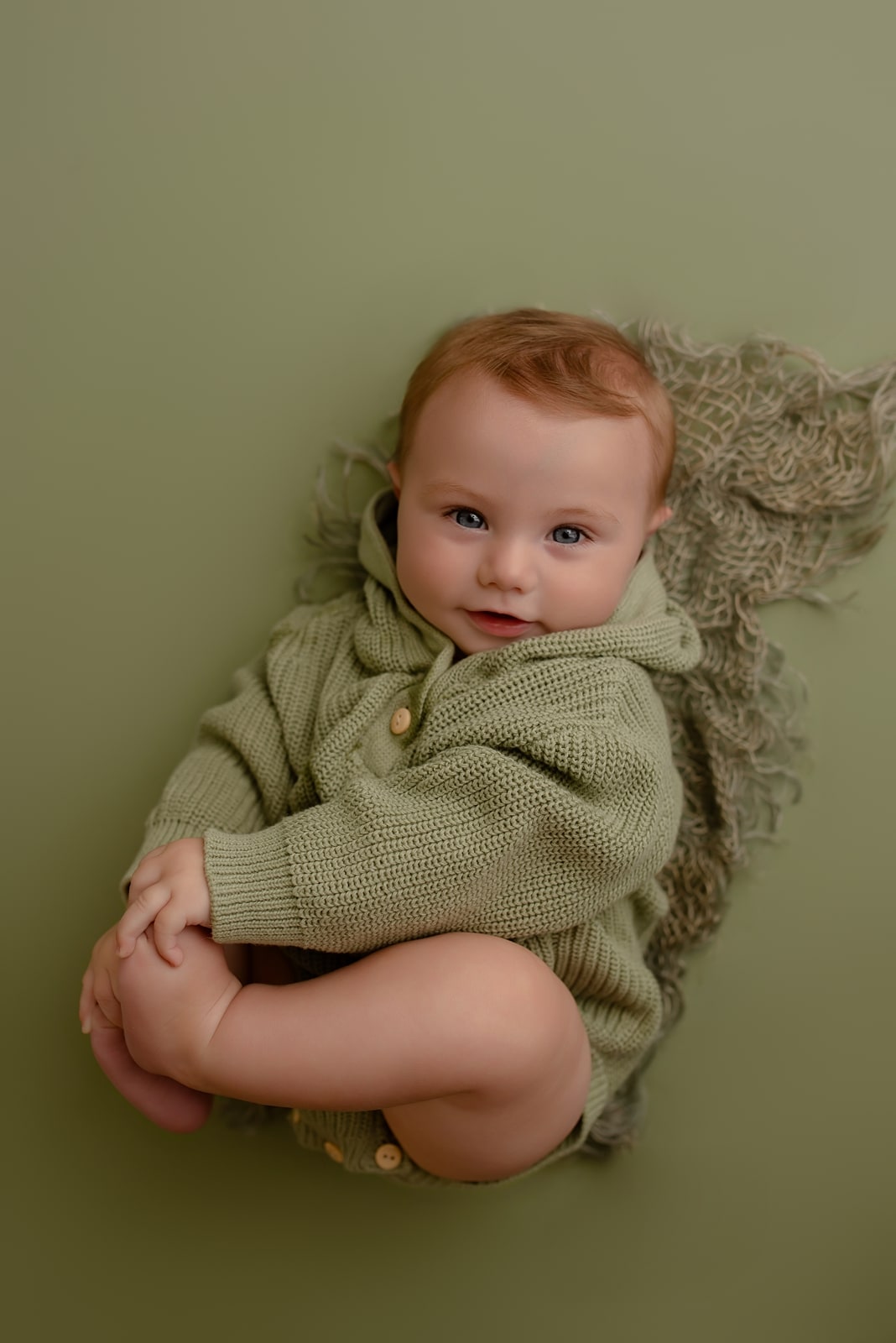 Baby in a green sweater curled up against a sage backdrop